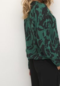 Blouse à motifs verts et noirs, coupe ample et manches longues, associée à un pantalon noir. Tissu doux et lisse avec un design fluide.
