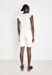 Haut créme à manches courtes avec des bords volantés, associé à un short beige clair jusqu'aux genoux avec un ourlet effiloché. Porté avec des sandales noires.