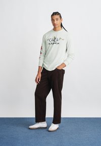 Camicia a maniche lunghe verde chiaro con grafiche di cartoni sul davanti e dettagli rossi sulle maniche, abbinata a pantaloni in velluto a coste marrone scuro e scarpe bianche.