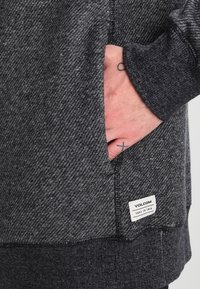 Szary, teksturowany hoodie z kieszenią z przodu, z naszywką z napisem "VOLCOM" oraz "TRUE TO THIS" w lewym dolnym rogu.