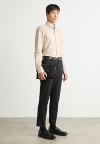 Helles beiges Button-Up-Hemd, schwarze Anzughose und Lederschuhe. Hält eine schwarze Leder-Clutch mit Klappe. Minimalistisches Design und taillierte Passform.