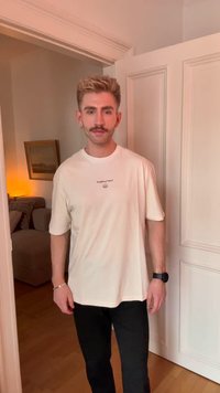 Homem com cabelo estilizado e bigode, vestindo uma t-shirt gráfica branca e calças pretas, em pé na entrada de uma sala suavemente iluminada.
