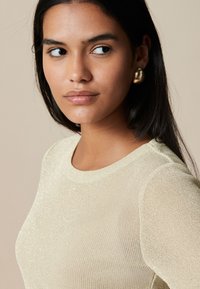 Goldfarbenes, strukturiertes Stricktop mit rundem Ausschnitt, das dünne, funkelnde Fäden aufweist. Glänzende goldene Ohrringe setzen Akzente im Look.