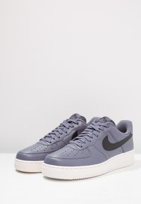 Sneakers Nike Air Force 1 w jasnofioletowej skórze, z czarnym logo, teksturowaną powierzchnią, okrągłym noskiem i białą gumową podeszwą.