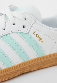 Zapatilla de cuero blanca con rayas verde menta, texto dorado "SAMBA" y suela de goma. Cuenta con cordones redondos y detalles de costura en zigzag.