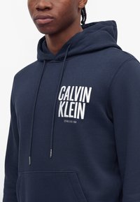 Jeune personne portant un sweat à capuche bleu marine avec "Calvin Klein Established 1988" imprimé en blanc sur la poitrine.
