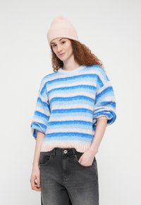 Νεαρή γυναίκα με σγουρά μαλλιά, φορώντας ροζ σκουφάκι, μπλε και λευκή ριγωτή μπλούζα και μαύρα τζιν, με το ένα χέρι στην τσέπη.