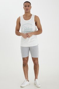 White cotton tank top with a "California" graphic, paired with light gray shorts. Model wears white sneakers, showcasing a casual outfit.