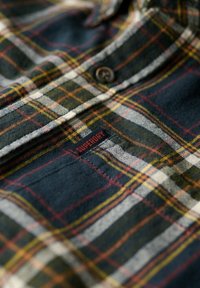 Chemise en flanelle de coton avec un motif à carreaux bleu foncé, vert et blanc, agrémentée d'accents rouges et d'une petite étiquette rouge "Superdry" près du bouton.