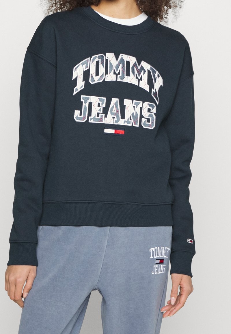 Námořnická modrá mikina s velkým bílým nápisem "TOMMY JEANS". Má švy s ribbed manžetami a lemem, měkkou tkaninu a volný střih.