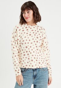 Pull en maille crème avec un motif à pois marron, col rond, poignets et ourlet côtelés, porté avec un jean bleu clair. Texture douce ; design décontracté.