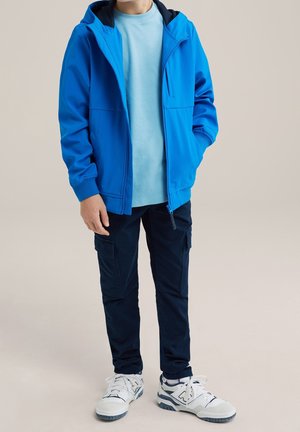 Enfant portant une veste zippée bleu vif, un t-shirt bleu clair, un pantalon cargo foncé et des baskets blanches, debout devant un fond uni.