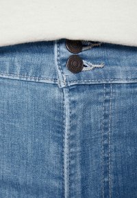Los jeans de mezclilla azul claro presentan dos botones marrón oscuro en la cintura, con costuras visibles y un ligero desgaste cerca de la parte superior.