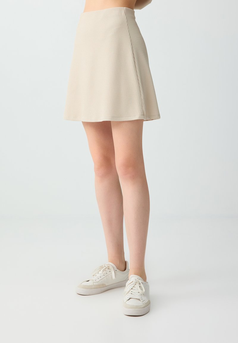 Femme portant une jupe beige côtelée en ligne A et des baskets blanches à lacets, debout devant un fond clair uni.