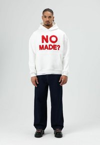 Witte hoodie met opvallende rode tekst "NO MADE?" op de voorkant, gecombineerd met donkerblauwe wijde jeans en zwarte schoenen.