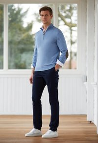 Himmelblauer Pullover mit Reißverschlusskragen und dunklen Ellenbogenflicken, kombiniert mit marineblauen Hosen und weißen Sneakers, auf einem Holzdeck platziert.