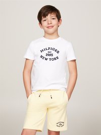 T-shirt blanche avec impression marine "HILFIGER EST. 1985 NEW YORK". Short jaune avec poche et détail de logo. Le modèle pose avec une main dans sa poche.