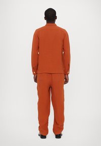 Set di camicia e pantaloni in lino color ruggine, caratterizzato da maniche lunghe, polsini con bottone e una vestibilità comoda. L'outfit è leggero e con una texture interessante.