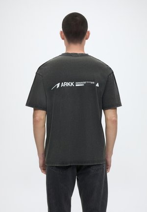 Άνδρας με κοντά μαλλιά που φοράει μαύρο μπλουζάκι με την επωνυμία ARKK και μαύρο τζιν, στέκεται κοιτώντας σε απλό λευκό φόντο.