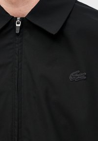 Gros plan d’une chemise noire zippée avec un col et un logo crocodile noir brodé sur la poitrine.