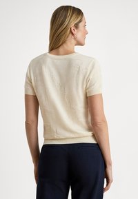 Krátky rukáv béžového svetra v textúrovanej pletenine, s okrúhlym výstrihom a rebrovaným lemom, v kombinácii s tmavomodrými nohavicami.