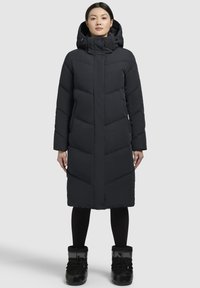 Sort pufjakke med høj krave, fuld lynlås og diagonalt quiltet mønster. Modellen bærer sorte snestøvler og leggings.