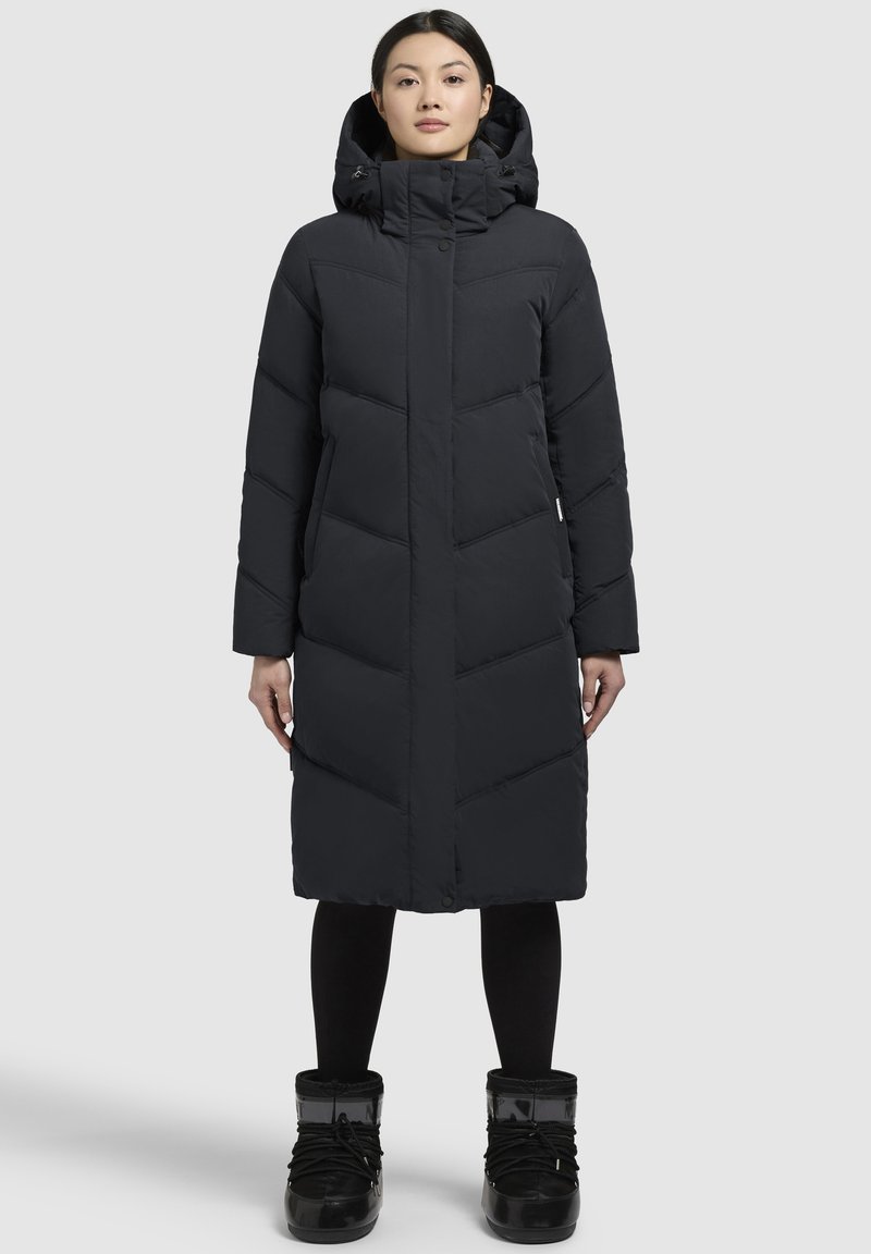 Sort pufjakke med høj krave, fuld lynlås og diagonalt quiltet mønster. Modellen bærer sorte snestøvler og leggings.