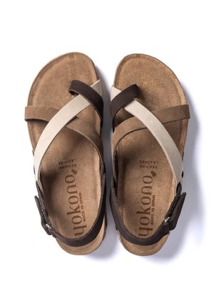 Een paar bruine suède sandalen met gekruiste beige en donkerbruine banden en verstelbare gespen op een witte achtergrond.