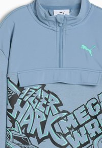 Felpa Puma di colore blu chiaro con zip a metà, logo ricamato e testo grafico audace "Tiger Shark" e "Mega Warrior" in nero e turchese.