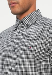 Schwarz-weiße karierte Hemdbluse mit Kragen, featuring dunklen Knöpfen und einem kleinen Logo auf der Brust.