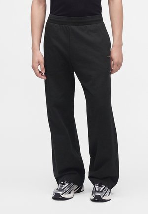 Personne portant un pantalon noir à jambes larges et des baskets argentées et noires, debout devant un fond clair uni.