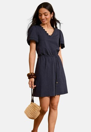 Navy Baumwollkleid mit einem gewellten Ausschnitt, kurzen Puffärmeln und tailliertem Schnitt, kombiniert mit einer geflochtenen Strohtasche.