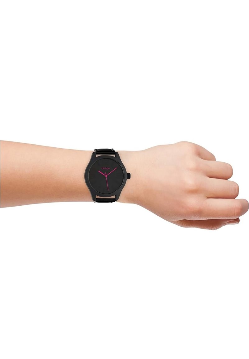 Montre analogique noire avec un cadran rond, marquages roses sur un bracelet texturé noir, portée sur un poignet à la peau claire contre un fond blanc.