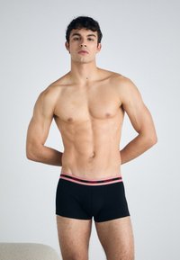 Boxeri negri cu o talie HUGO roz, având un design ajustat și o textură netedă din bumbac. Modelul stă cu încredere, cu brațele pe lângă corp.