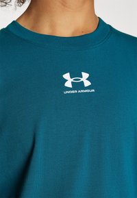 Person trägt ein türkisfarbenes Sweatshirt mit rundem Ausschnitt und weißem Under Armour-Logo, das zentriert unter dem Halsausschnitt angebracht ist.