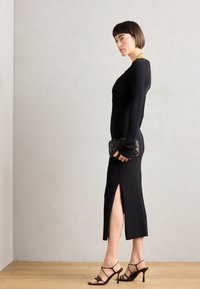 Rochie neagră din tricot cu nervuri, având o fanta laterală, asortată cu pantofi cu toc subțiri negri și o geantă texturată neagră. Textură netedă, design slim fit.