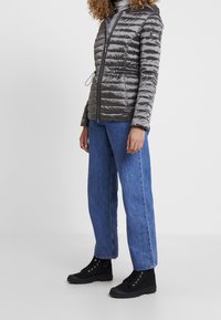 Graue Steppjacke mit durchgehendem Reißverschluss, horizontaler Stepptextur, verstellbarem Kordelzug in der Taille, getragen mit blauen weiten Jeans und schwarzen Stiefeln.