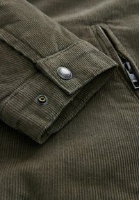 Tejido de corduroy verde oliva con textura acanalada, que presenta un detalle de cierre con botón a presión y cremallera en el lado izquierdo.