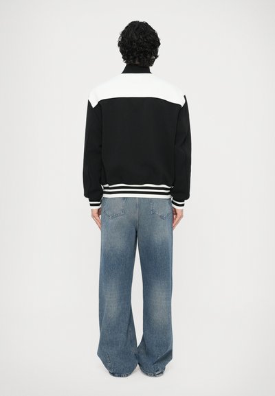 Čierno-biela bomber bunda s vysokým golierom, pruhovanými manžetami a voľným strihom, v kombinácii so svetlomodrými džínsami s širokými nohavicami.