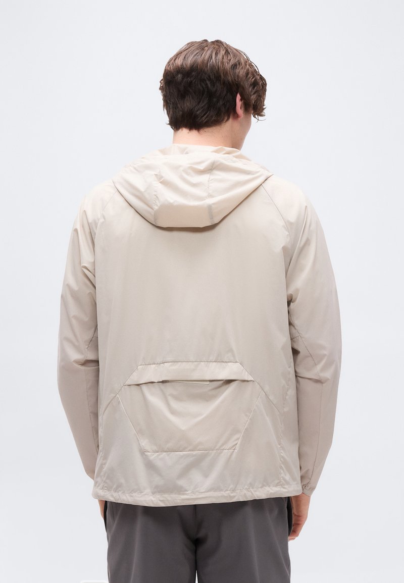 Homme portant une veste à capuche beige avec une poche arrière, debout de dos sur un fond blanc uni.