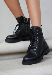 Bottes de cheville en cuir noir avec un bout rond, fermeture à lacets à l'avant, œillets en métal et zip sur le côté. Semelle en caoutchouc épaisse avec un motif de semelle pour une meilleure adhérence.