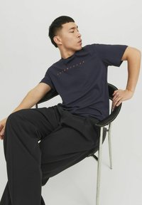 Navyblaues T-Shirt mit orangefarbenem Logo-Text, aus Baumwolle, Rundhalsausschnitt, kurze Ärmel, kombiniert mit lockeren schwarzen Hosen, sitzend auf einem Stuhl.