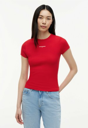 Woman wearing fitted red short-sleeve shirt with "tommy jeans" logo and light blue jeans, standing with one hand behind her back.