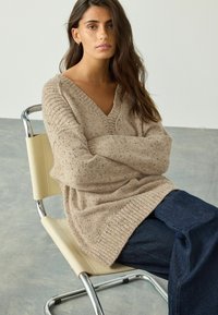 Beige striktrøje med V-hals, tekstureret garn og ribbede manchetter, parret med mørke denimjeans. Model sidder på en stol i krom og beige.