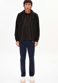 Felpa nera con zip e tasche a marsupio, indossata sopra una t-shirt nera coordinata. Abbinata a pantaloni blu navy e sneaker bianche.