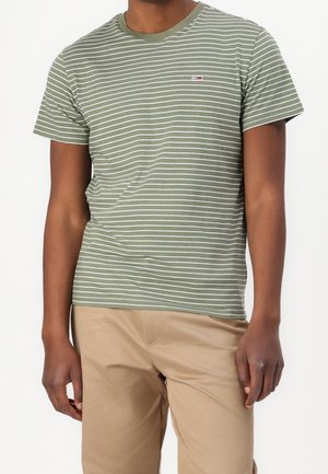 Mężczyzna w oliwkowo-białej, paskowanej koszulce z krótkim rękawem i beżowych spodniach na jednolitym, białym tle.