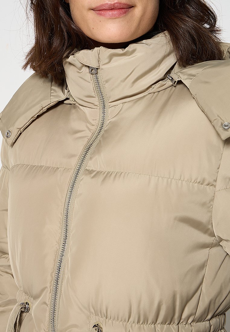 Doudoune beige avec un col haut, fermeture zippée double et accents d'épaule boutonnés. Matériau lisse et léger avec une texture matelassée.