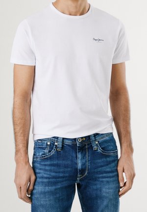 Männlicher Torso, der ein weißes kurzärmeliges Rundhals-T-Shirt mit kleinem Textlogo auf der Brust und blaue Jeans trägt.