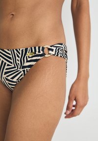 Bikini apakšda ir ar ģeometrisku melnu un bēšu rakstu, sānu gredzenu apdari un gludu audumu ar maigu tekstūru.