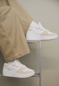 Hvite og kremfargede sneakers med glatt skinnøvre, snøring, perforert tå og opphøyd såle, kombinert med beige bukser.